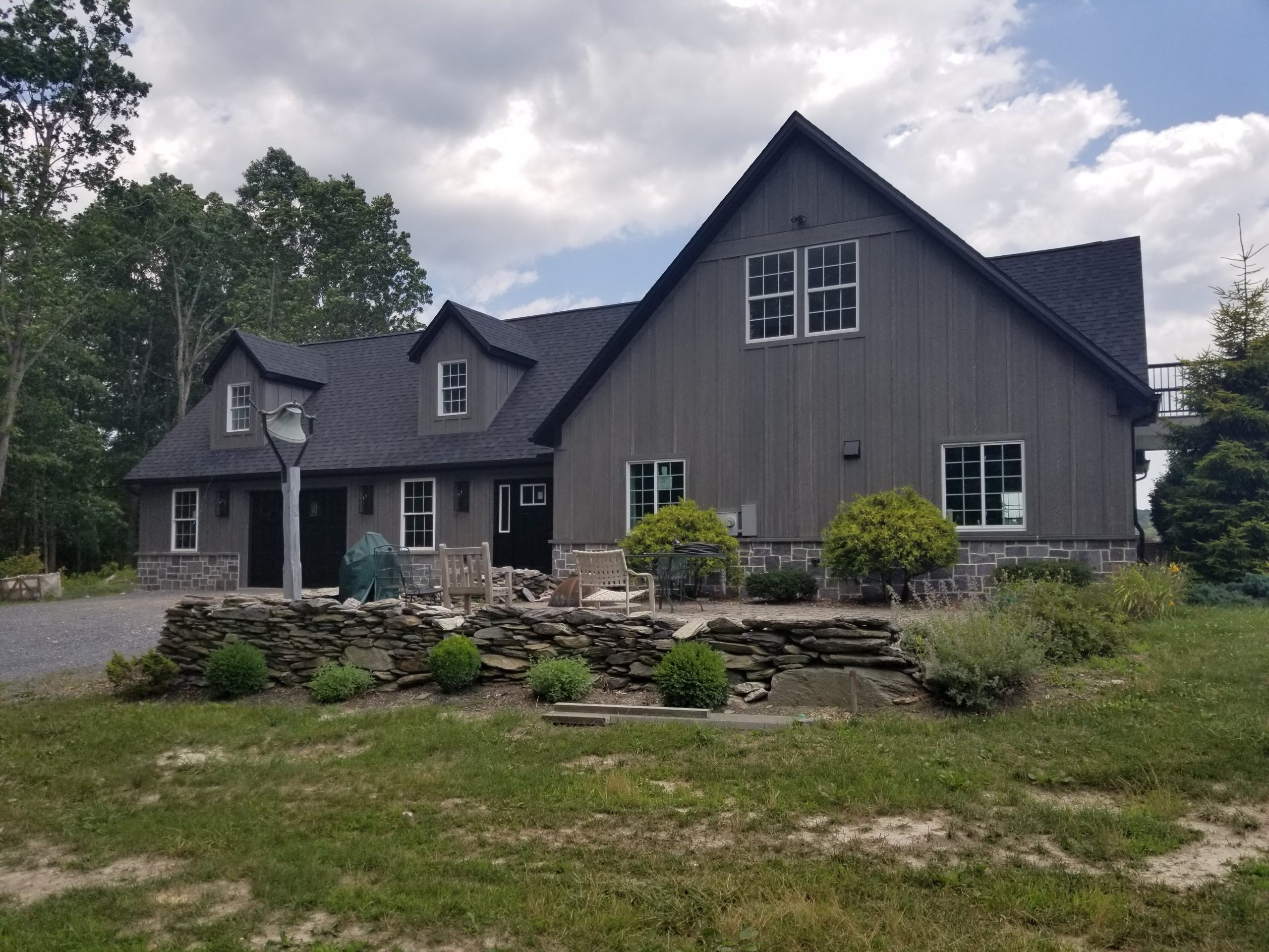 Back exterior view of a remodeled home with stone accents, gray siding, black roofing, an attached 1 car garage, and a patio with chairs.