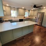 Overview of a renovated kitchen with sage green cabinets, white counter tops, white walls, dark brown hardwood flooring, a stainless steel fridge, a black ceiling fan, and a white farmhouse sink.