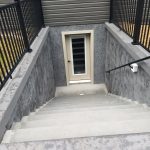 Close up of stairs leading down to a basement entrance with a beige door and large window.