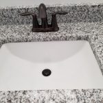 Close up of a dark metallic sink faucet, white and gray granite countertop, and white sink.