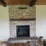 Close up of a remodeled electric stone fireplace with a wood shelf and overhead lighting.