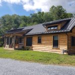 Front and right side exterior view of a custom home with rustic wood siding, black windows, black metal roofing, and a front porch with log posts.