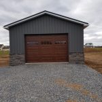 Front exterior view of a pole shed with dark gray siding, a brown garage door, and stone siding accents.