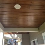 Close up of a porch ceiling with a brown wood surface, white trim, and a round white light.