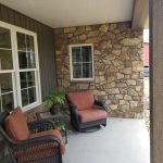 Close up of a new home's covered front porch with 2 brown and orange wicker arm chairs, a stone accent wall, and brown siding.