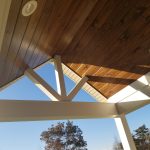 Close up of a porch ceiling with a brown wood surface, white trim, white beams, and a round white light.