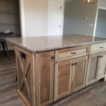 Close up of a kitchen island made with stained brown wood and a speckled granite countertop in a kitchen with light gray walls and hardwood flooring.