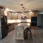 Overview of a new kitchen with brown stained cabinets, speckled granite countertops, a kitchen island with 3 black stools, dark stainless steel appliances. light colored walls, and hardwood flooring.