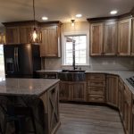 Overview of a new kitchen with brown stained cabinets, speckled granite countertops, a kitchen island with 3 black stools, dark stainless steel appliances. light colored walls, and hardwood flooring.