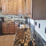 Kitchen with black and gold quartz countertops, brown cabinets, a stainless steel double sink, white tile backsplash, and light wood flooring.