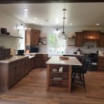 Remodeled kitchen with a brown and white island with black stool chairs, wood flooring, white tile backsplash, brown cabinets, and white marble countertops.