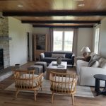 Remodeled living room with wood ceiling and exposed beams, a gray stone fireplace, wood flooring, 2 couches, and 2 wood armchairs.