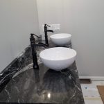 Close up of a sink vanity with black marble countertop, white bowl sink basins, and black sink faucets.