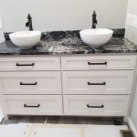 Close up of a white sink vanity with black marble countertop, white bowl sink basins, and black sink faucets.