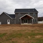 Back exterior view of a new home with dark gray siding, white trim, a glass sliding door, black roofing, and a back porch with an a-frame roof.