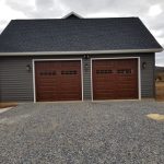 2-car garage with dark gray siding, 2 brown garage doors, black roofing, white trim, and outdoor lights.