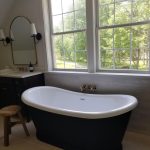 Close up of a soaking tub and wooden stool next to 2 sink vanities in a remodeled bathroom with large windows overlooking the home's yard.
