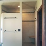 Outside view of a tile shower with light colored tile walls, 2 patterned accents strips, a shower hook, 2 towel racks, gray tile flooring, and a built-in corner shower seat.
