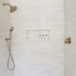 Close up of a new shower in a remodeled bathroom with white tile walls, white tile floors, built in wall shelves, and 2 metallic shower heads.