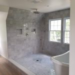 Close up of a shower and tub combination with a white soaking tub, gray tiled walls, and gray tiled shower flooring in a remodeled bathroom with 2 silver shower heads.