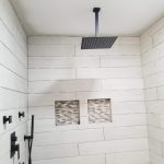 Close up of a white tile shower with black shower faucets, a metallic shower head, and beige-colored tile built-in shelving.