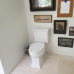 White toilet in a corner with white walls, tan tile flooring, and an assortment of vintage ship framed wall art.