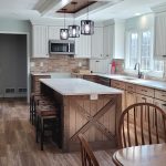 Remodeled kitchen with wood flooring, rustic wood cabinets, a kitchen island with 4 brown stools, white countertops, exposed beams, and 3 pendant lights over the kitchen island.