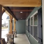 Front porch after remodel with wood plank ceiling, stone pillar accents, green vinyl siding, exposed wood beams, and white windows with black shutters.