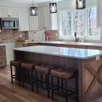 Remodeled kitchen with rustic wood cabinets, wood flooring, an island with 4 stools, brown tile backsplash, 3 pendants lights, and white countertops.
