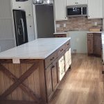 Remodeled kitchen with rustic wood cabinets, wood flooring, a kitchen island, brown tile backsplash, and white countertops.