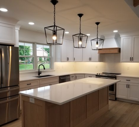 New kitchen with plank flooring, white walls, white cabinets, white countertops, a wood paneled kitchen island, stainless steel appliances, a black sink, plank flooring, and 3 black hanging lights above the island.