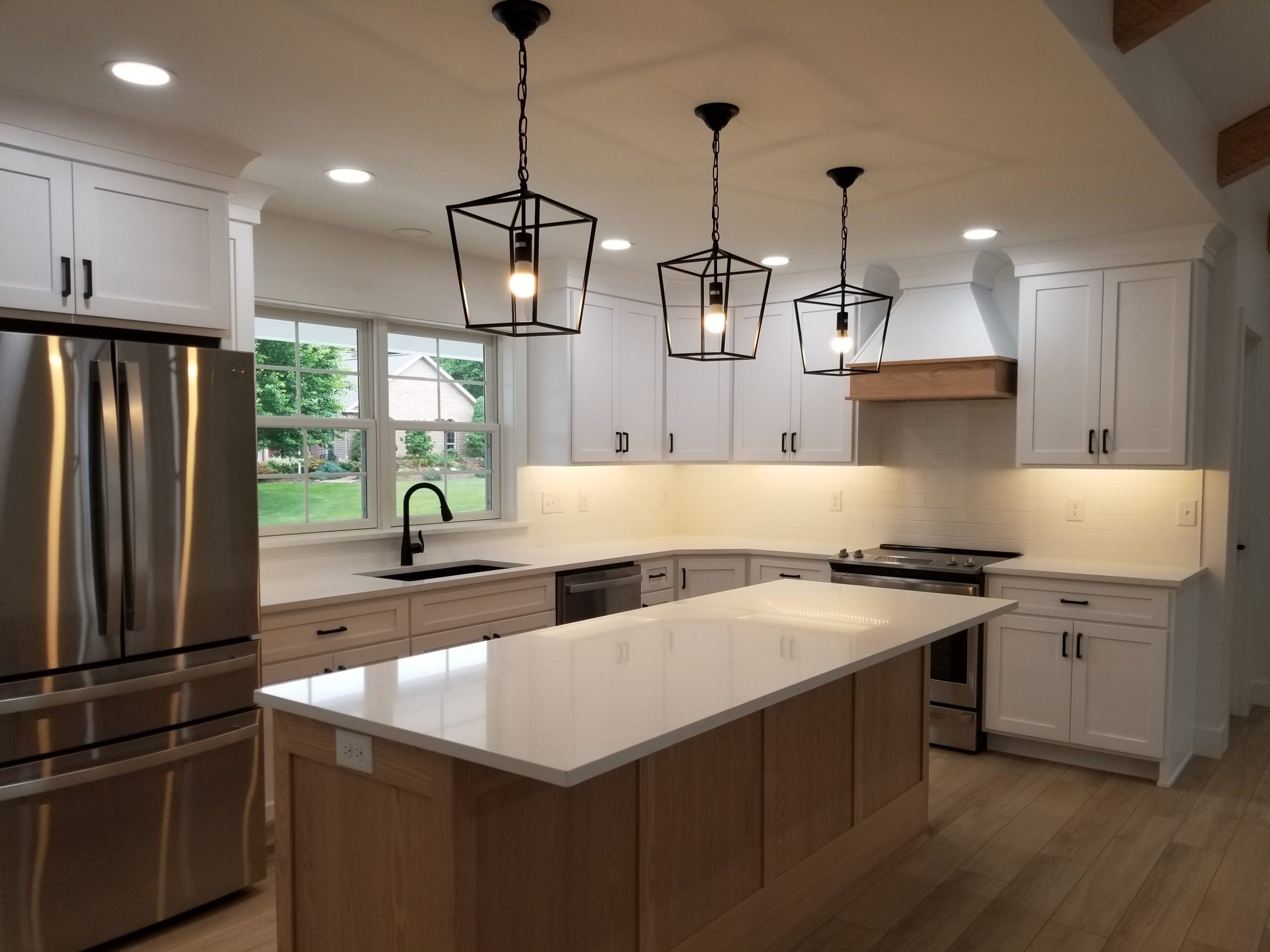 New kitchen with plank flooring, white walls, white cabinets, white countertops, a wood paneled kitchen island, stainless steel appliances, a black sink, plank flooring, and 3 black hanging lights above the island.