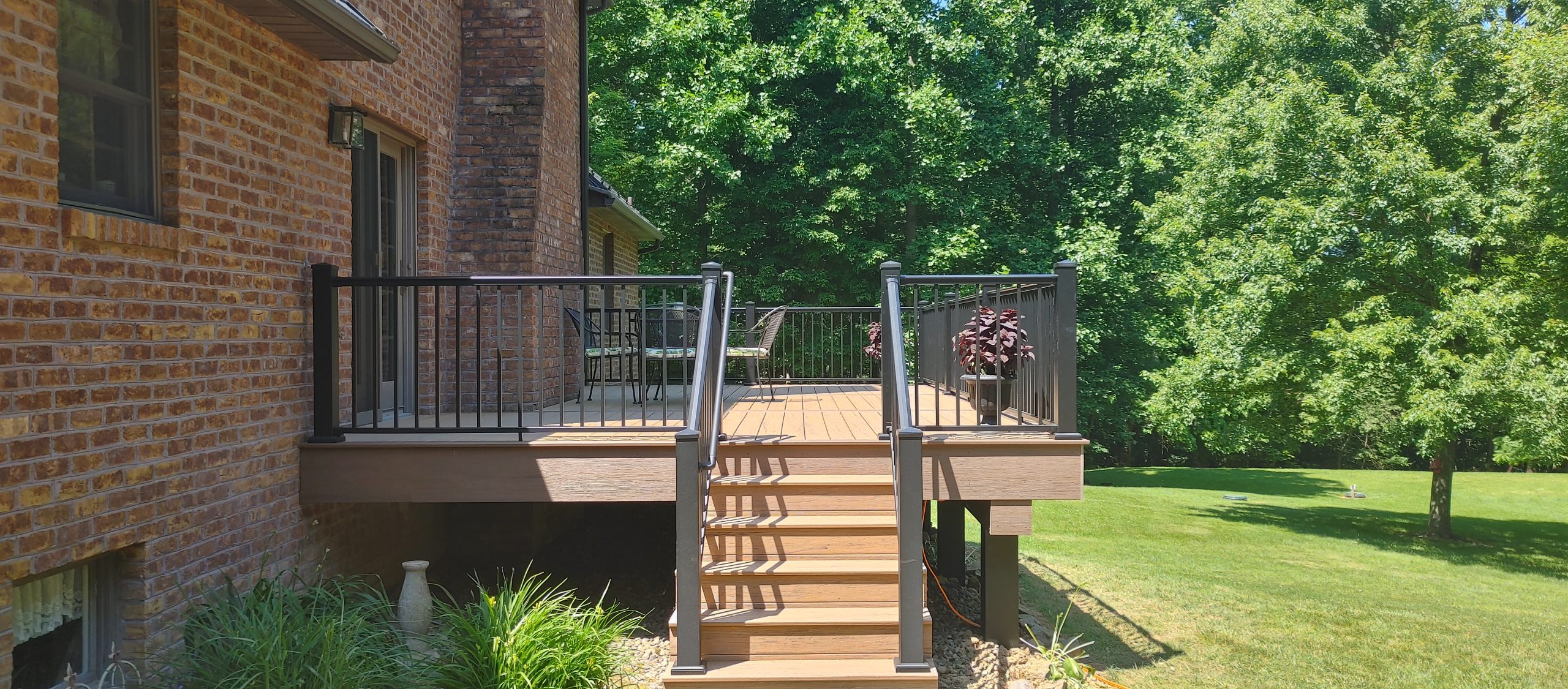 New, tan deck with black railings attached to a red brick house in a backyard surrounded by trees.