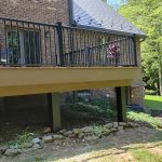 New, tan deck with black railings attached to a red brick house in a backyard surrounded by trees.