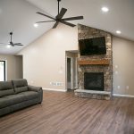 Living room with tan walls, hardwood floors, a gray leather couch, 2 ceiling fans, and a large stone fireplace with a mounted tv.