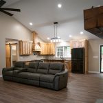 Interior of a home with tan walls, hardwood floors, a white ceiling, a large gray leather couch, and a kitchen with natural wood cabinets.