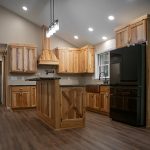 A kitchen with natural wood cabinets, a natural wood kitchen island, hardwood flooring, tan walls, and a large black fridge.