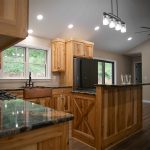 A kitchen with natural wood cabinets, dark quartz countertops, a natural wood kitchen island, hardwood flooring, tan walls, and a black fridge.