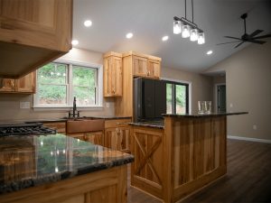 A kitchen with natural wood cabinets, dark quartz countertops, a natural wood kitchen island, hardwood flooring, tan walls, and a black fridge.