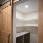 Natural wood sliding barn door opening into a kitchen pantry with white shelves, tan walls, and a white quartz surface.