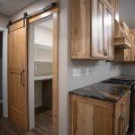 Kitchen with natural wood cabinets and black quartz countertops next to a sliding barn door leading into a kitchen pantry with white shelves.