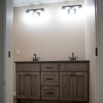 Large bathroom vanity with 2 sinks, 3 drawers, and 2 sets of cabinets inside a bathroom with tan walls, brown hardwood flooring, and 2 mounted lights above the sinks.