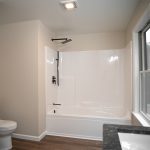 Bathroom with tan walls, brown hardwood flooring, a white shower with a bathtub, a white toilet, and a white window.