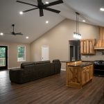 Interior of a home with tan walls, hardwood floors, a white ceiling, a large gray leather couch, 2 ceiling fans, a kitchen with natural wood cabinets, a natural wood kitchen island, and a set of glass doors leading out to a deck.