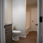 White door leading into a bathroom with tan walls, a brown wood sink vanity, a white toilet, and a white shower with a bathtub,