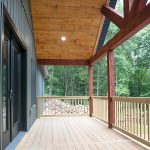 A covered wood deck with red posts and beams, natural wood railings, a natural wood ceiling, and round ceiling lights attached to a home with green siding.