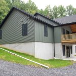 Front and side exterior view of a 2-story home with a white first story, green second story, black windows, black asphalt roofing, and a second story covered wood deck.