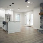 Combined kitchen and living area with light gray walls, vinyl plank flooring, a white kitchen island with a black sink faucet, white kitchen cabinets, and a large stone fireplace with a wood beam shelf.