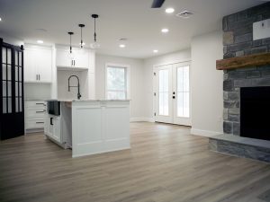 Combined kitchen and living area with light gray walls, vinyl plank flooring, a white kitchen island with a black sink faucet, white kitchen cabinets, and a large stone fireplace with a wood beam shelf.