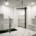 Kitchen with light colored walls, white cabinets, white marble countertops, and a black sliding door leading into a white pantry.